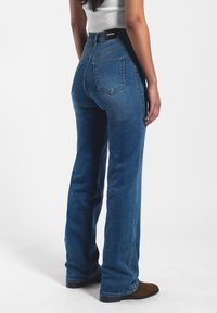 Jeans de mezclilla azul oscuro acampanados con una cintura alta, que cuentan con dos bolsillos traseros, costuras visibles y un parche de logo similar al cuero en la cinturilla.