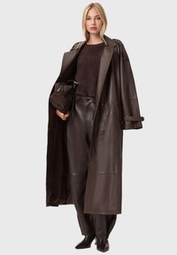 Brązowy skórzany płaszcz z dużymi guzikami, zestawiony z ciemnobrązowym swetrem i skórzanymi spodniami. Modelka trzyma torebkę o dopasowanej fakturze.