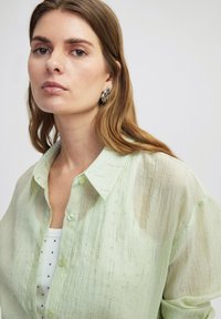 Camisa de botones semitransparente en verde claro con un patrón texturizado y sutiles acentos metálicos, llevada sobre una blusa blanca con puntos.