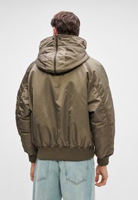 Mężczyzna w oliwkowej, pikowanej kurtce z kapturem i jasnoniebieskich dżinsach, stojący tyłem do kamery na jednolitym tle.