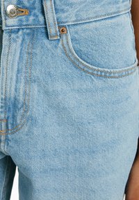 Helle blaue Jeans aus Denim mit hohem Bund, Kupferriegeln und kontrastierender Naht. Enthält Fronttaschen und eine strukturierte Oberfläche.
