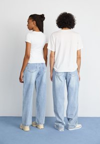 To personer står med ryggen til, iført hvite topper og lyseblå jeans. Jeansene har vide ben og en falmet tekstur.