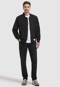 Schwarze Bomberjacke, leichtes Material, Reißverschluss, gerippte Bündchen, mit Reißverschlusstaschen, über einem weißen T-Shirt und schwarzen Jeans getragen, mit gemusterten Schuhen.