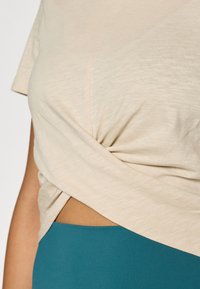 Ανοιχτό μπεζ τοπ με κοντά μανίκια και κόμπο στη μέση, σε συνδυασμό με ψηλόμεσο πετρόλ κάτω μέρος, που αναδεικνύει μια ομαλή υφή.