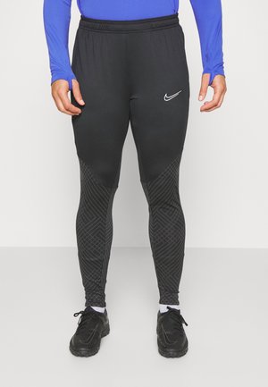 Homme portant un pantalon de sport noir Nike avec des motifs géométriques sur le bas des jambes, des chaussures de course noires et un haut bleu à manches longues.