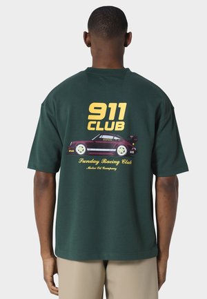 Homme portant un t-shirt vert foncé avec le texte jaune "911 CLUB" et une illustration de voiture de course vintage dans le dos, debout devant un fond uni.