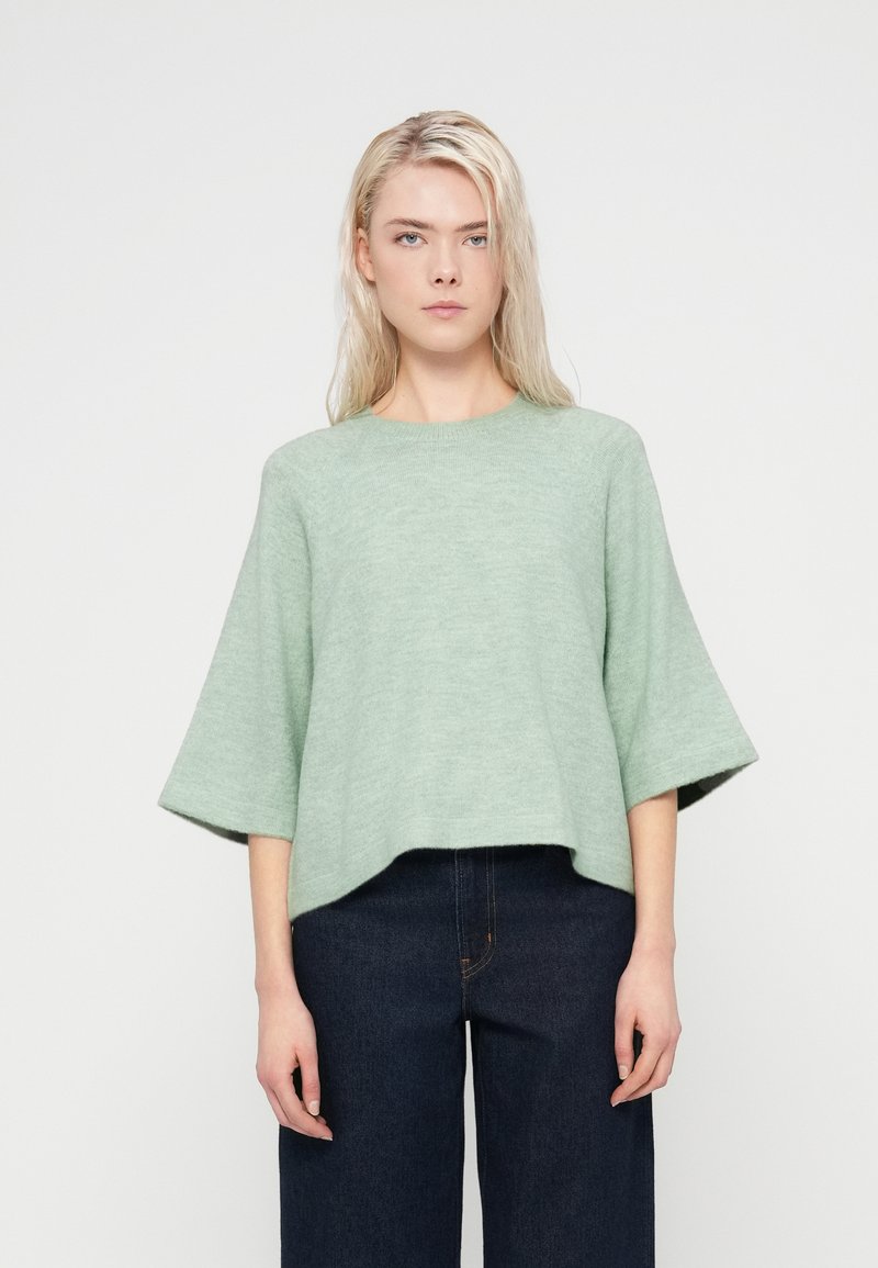 Pull ample vert clair avec un col rond et des manches trois-quarts. Texture douce avec un motif tricoté subtil. Associé à un jean foncé.