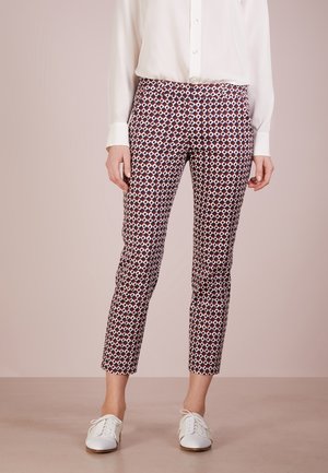 Kobieta ubrana w białą koszulę na guziki, wzorzyste wąskie spodnie cropped z geometrycznym nadrukiem oraz białe sznurowane buty, stojąca na jednolitym podłożu.