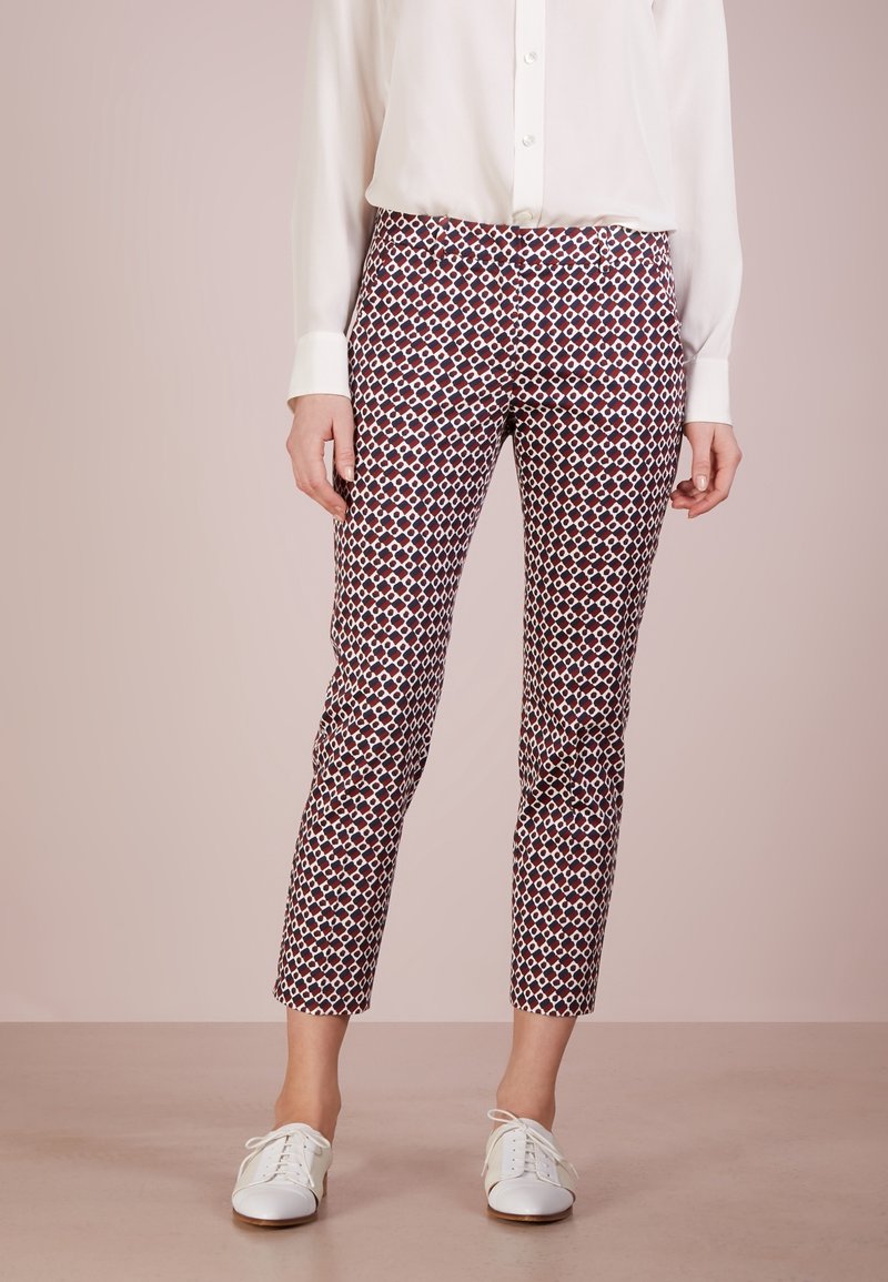 Femme vêtue d'une chemise blanche à boutons, pantalon slim cropé à motifs géométriques, et chaussures blanches à lacets, debout sur un sol uni.
