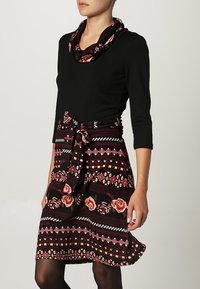 Top lungo nero a maniche lunghe con scollatura drappeggiata abbinato a una gonna borgogna con motivi floreali e geometrici, legata in vita.