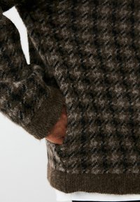 Brūns un pelēks rakstains adīts džemperis ar teksturētu apdari, kurā ir redzama roka sānu kabatā un ribota apakšmala un piedurknes.