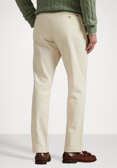 Μπεζ βαμβακερό παντελόνι με ίσια γραμμή, διαθέτει πίσω τσέπες με κουμπώματα. Φοριέται με ένα πράσινο υφασμάτινο πουλόβερ.