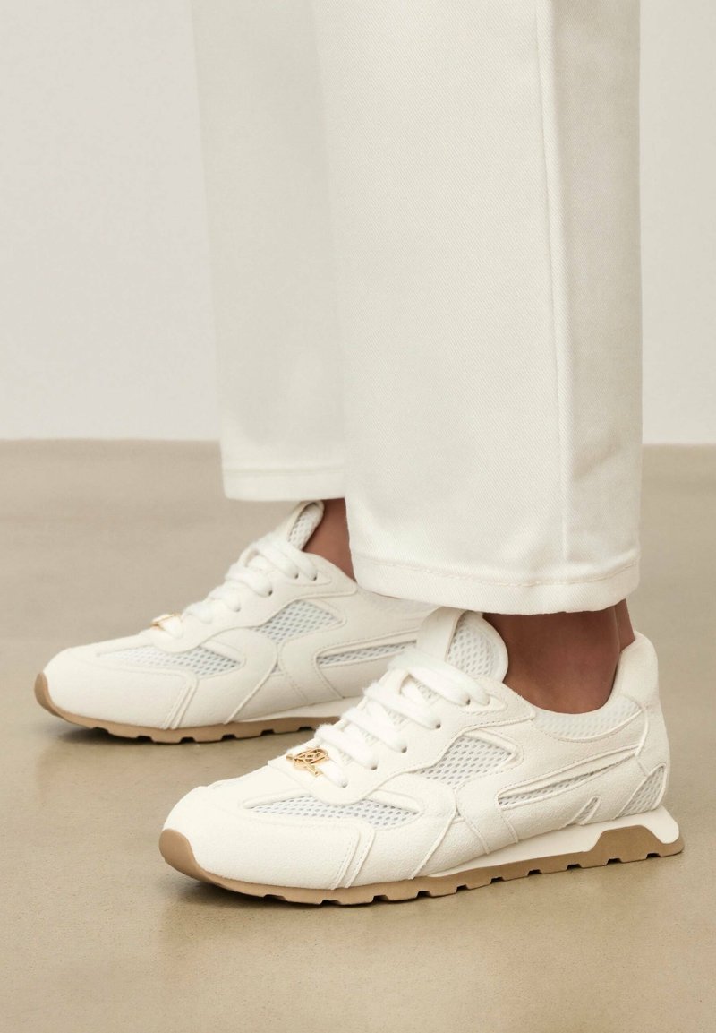 Baskets en maille blanche et cuir avec semelles beiges portées avec un pantalon blanc sur un sol neutre, montrant des vues latérales et avant des chaussures.