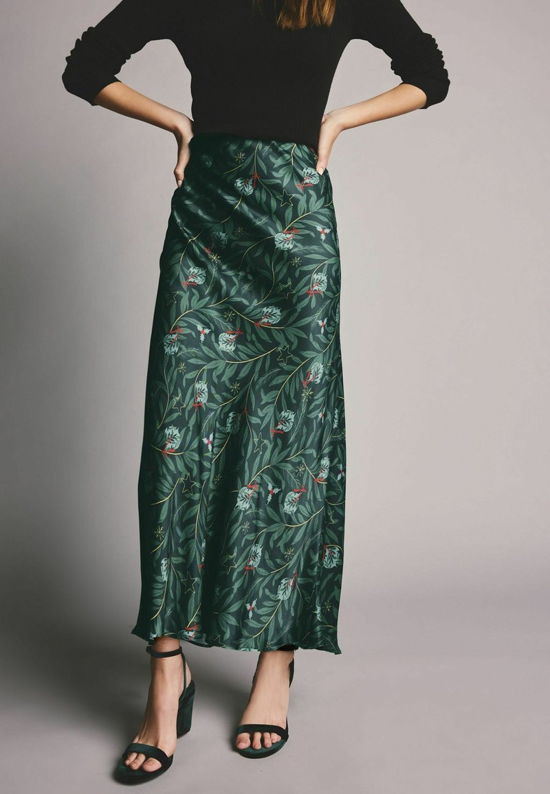 Jupe longue en satin vert avec motif floral et feuilles, longueur mi-mollet, texture lisse, portée avec des talons ouverts noirs.