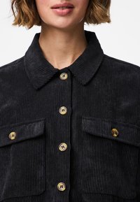 Žena odjevena u crnu kordonnu jaknu s velikim dugmadima u obliku kornjačevine i dva džepa na prsima, prikazana od ramena do usana.