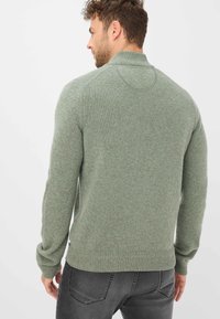 Olivgrüner Strickpullover mit hohem Kragen, gerippten Bündchen und Saum. Strukturiert stoff, taillierter Schnitt. Graue Jeans sind am unteren Rand sichtbar.