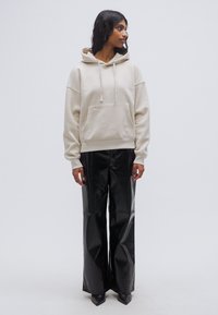 Κρεμ φούτερ με κουκούλα, μπροστινή τσέπη και κορδόνια, σε συνδυασμό με μαύρο γυαλιστερό παντελόνι φαρδύ με ψηλή μέση και μυτερά μαύρα παπούτσια.