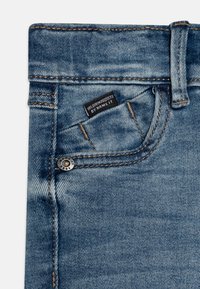 Jeans de mezclilla azul claro que cuentan con un cierre de cremallera, trabillas para el cinturón y un pequeño bolsillo frontal con una etiqueta de marca cosida en la cinturilla.