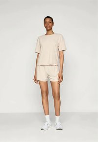 Bež majica s teksturom i kratkim rukavima, kombinirana s odgovarajućim šortsevima. Oba imaju opušteni kroj, upotpunjeni bijelim sportskim čarapama i cipelama.