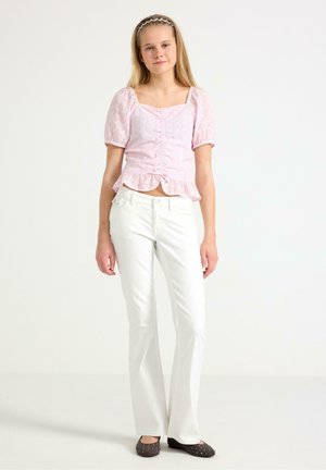 Ung kvinde iført en lyserød rynket bluse, hvide bukser og sorte flats med nitter, står foran en ensfarvet hvid baggrund.