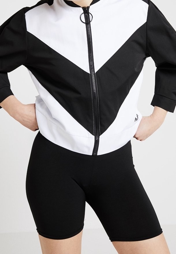 Svart och vit zip-up jacka med framträdande chevronmönster; matchad med svarta, stretchiga shorts. Båda är tillverkade av ett slätt, atletiskt tyg.
