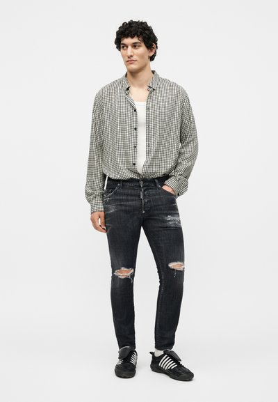 Νέος άνδρας που φοράει ασπρόμαυρο καρό πουκάμισο πάνω από λευκό τοπ, σκισμένο μαύρο στενό τζιν και μαύρα αθλητικά παπούτσια, στέκεται με το ένα χέρι στην τσέπη.
