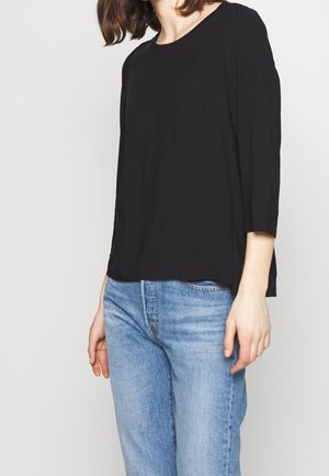 Kvinde iført en sort langærmet top og lyseblå jeans, stående mod en ensfarvet lys baggrund.