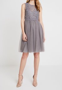 Robe cocktail grise sans manches, avec un corsage en dentelle et une jupe en tulle. Présente un motif floral sur la partie supérieure et une texture légère.