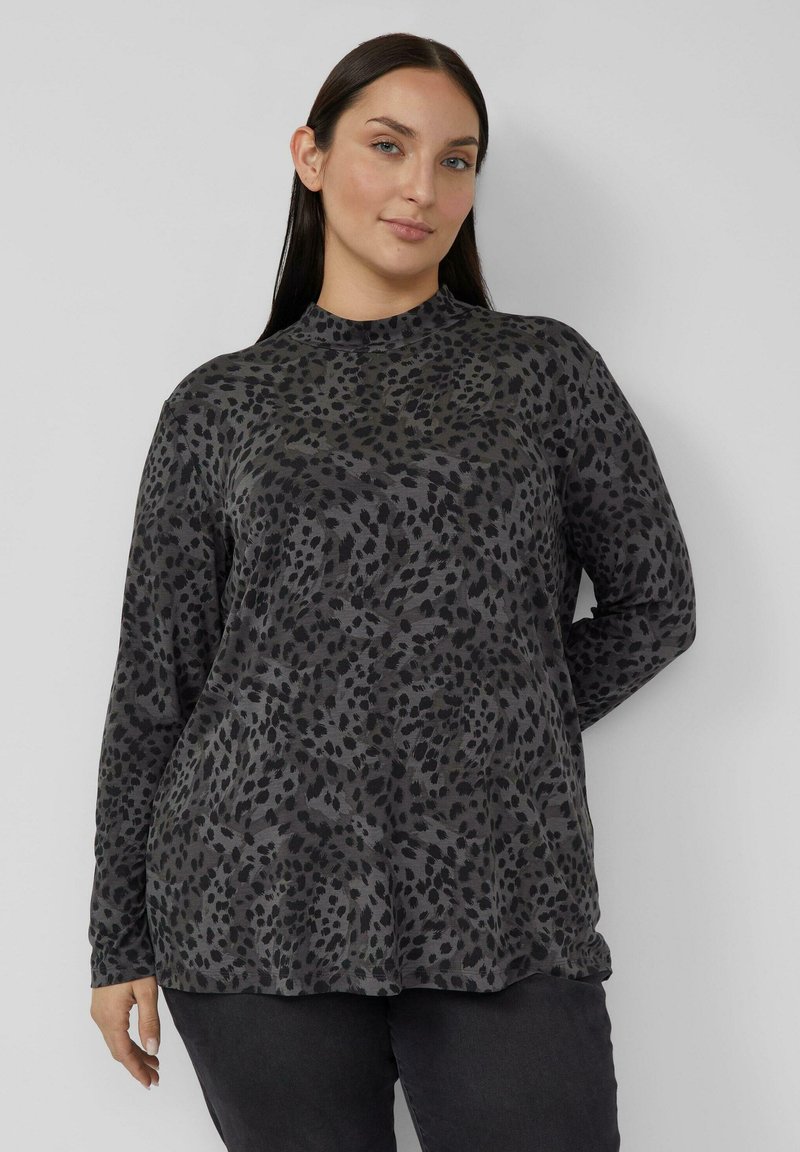 Kobieta o długich ciemnych włosach, ubrana w szarą bluzkę w czarne plamy z długim rękawem i ciemne spodnie, stojąca na jasnym, jednolitym tle.