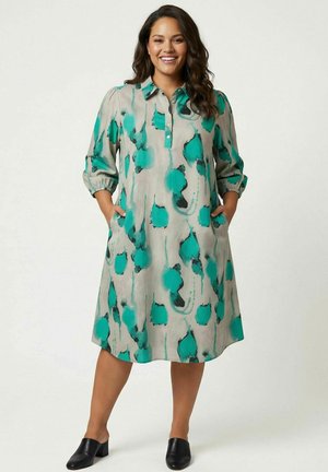 Frau trägt ein knielanges, geknöpftes Langarmkleid mit blau-grünem abstraktem Muster, steht mit den Händen in den Taschen und lächelt.