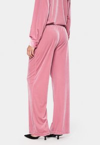 Rozā samta platās bikses ar mīkstu tekstūru, ar elastīgu jostu un smalku šuvju detaļām. Velkotas ar melniem norādītiem apaviem.