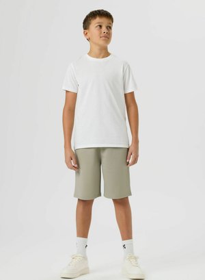 Garçon debout, portant un t-shirt blanc uni, un short beige au genou, des chaussettes blanches et des baskets blanches, regardant légèrement vers le haut.