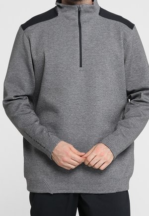 Homme portant un pull gris à manches longues avec un col zippé noir au quart et des panneaux noirs aux épaules, mains jointes devant.