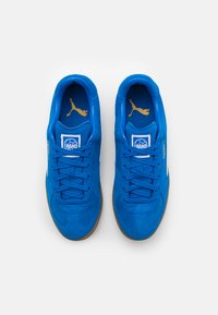 Blauwe suède sportschoenen met een rubberen gumzool, voorzien van blauwe veters, gouden logo-accenten en een witte zijstreep.
