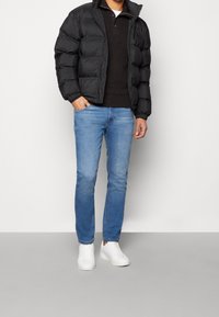 Man som bär en svart puffjacka, svart krage-sweater, ljusblå jeans och vita sneakers står med ena handen i fickan.