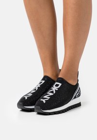 Nohy v černých slip-on teniskách DKNY s bílým logem, texturovaným pleteným svrškem, bílou střední podešví a černou vroubkovanou spodní podrážkou na bílém pozadí.