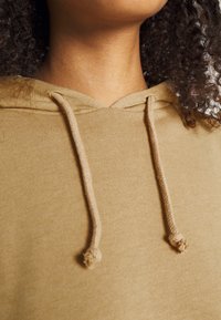 Sudadera de color marrón claro con una textura suave, que cuenta con un cierre de cuello ajustable mediante cordón y material acanalado. Vista en primer plano de la zona del cuello.