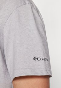Šedé tričko s krátkým rukávem s hladkým povrchem a černým logem "Columbia" na levém rukávu.