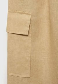 Beige linnen stof met een detail van een opgestikte zak. De textuur is glad met een licht kreukelige uitstraling, wat het natuurlijke materiaal benadrukt.