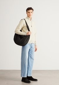 Fekete hobo táska, sima anyagból, vállon viselve. A modell bézs cipzáros dzsekiben, világos kék farmernadrágban és fekete csizmában.