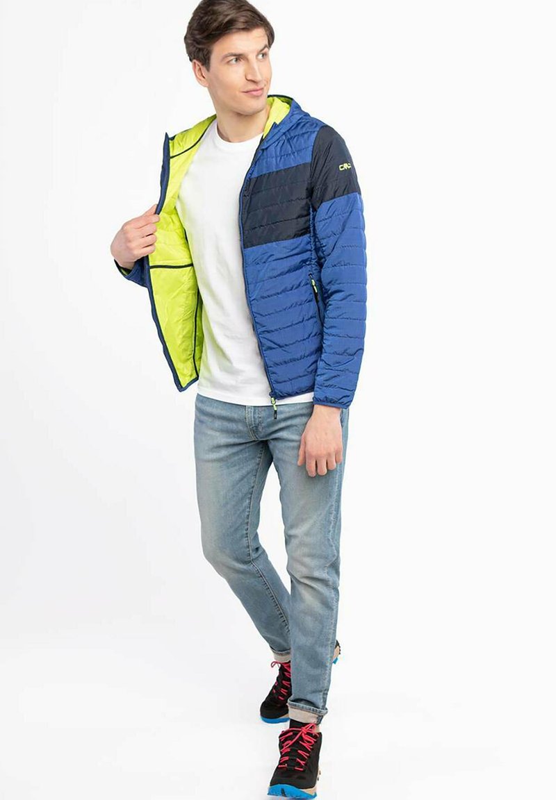 Gepolsterte Jacke in Marineblau und Schwarz, mit einem leuchtend gelben Innenfutter und Akzenten. Verfügt über eine Kapuze und Reißverschlusstaschen. Blaue Jeans und schwarze Sneakers.