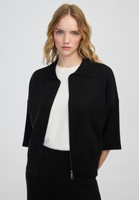 Žena s vlnitými blond vlasy, oblečená v černém svetr s zipem přes bílou košili a černé kalhoty, stojí před jednolitým světlým pozadím.