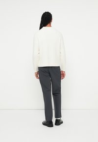 Maglione crema con scollatura rotonda e orlo a coste, abbinato a pantaloni grigi scuri a sigaretta. Modella vista da dietro con capelli intrecciati.