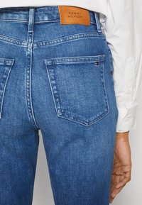 Modré džíny z denimu s vysokým pasem, které mají hnědou koženou etiketu na pase a zadní kapsy s detailem šití. Hladká textura.
