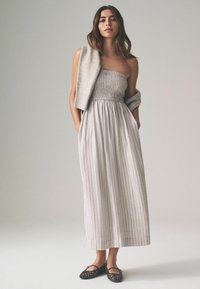 Strapless, gerimpelde maxi jurk in lichtgrijs met verticale strepen; gecombineerd met een grijze gebreide cardigan over één schouder en zwarte flats.