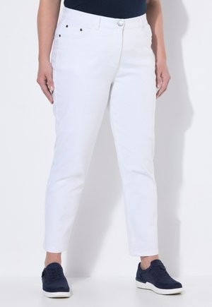 Person trägt weiße, schlanke Hose, marineblaue Turnschuhe und ein marineblaues Oberteil, steht vor einem weißen Hintergrund.