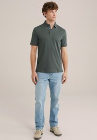 Polo de color verde oscuro con mangas cortas y cuello, combinado con unos vaqueros claros y zapatillas beis. Diseño sencillo y ajustado.