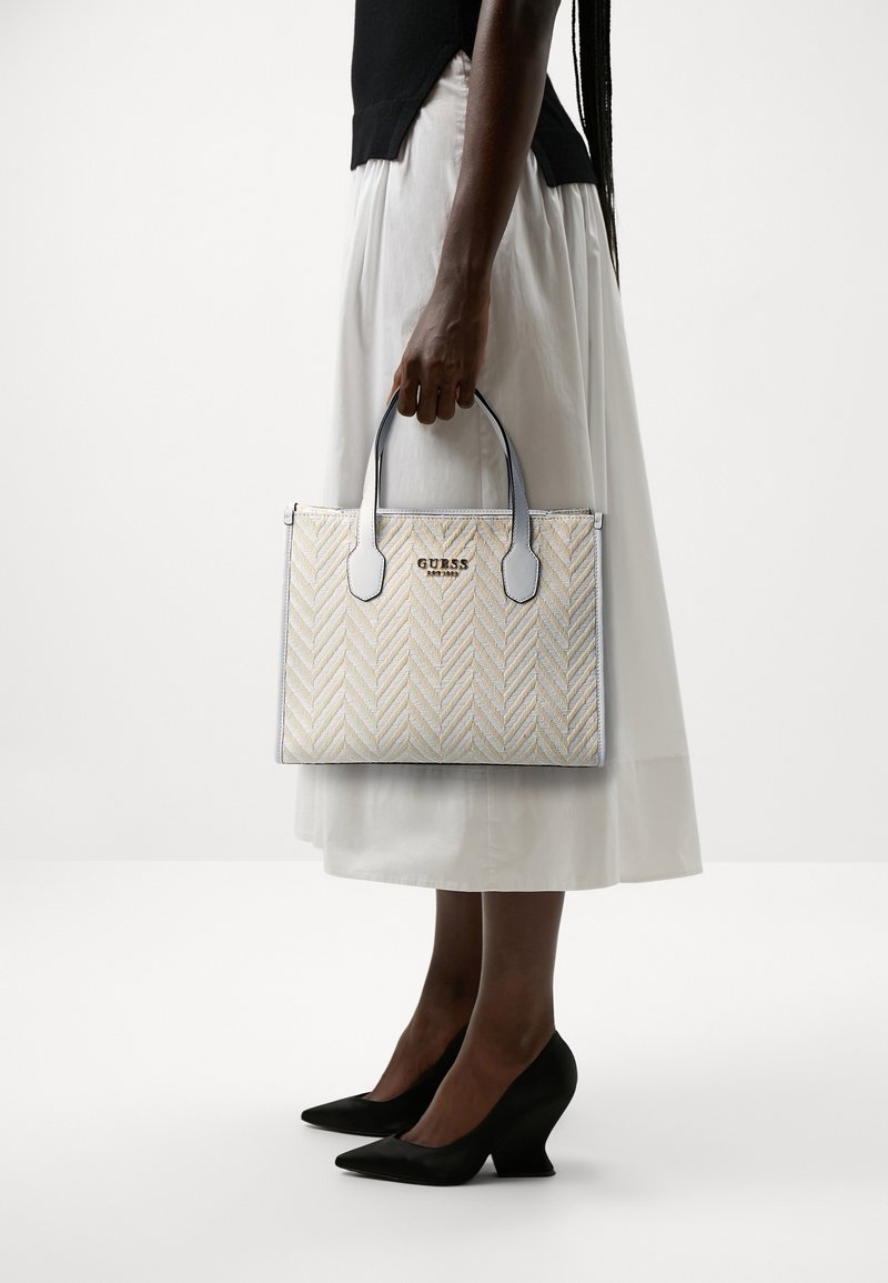 Femme portant un haut noir, une jupe blanche, des talons noirs tenant un sac à main blanc à motifs Guess avec des poignées noires sur le côté.