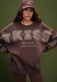 Felpa marrone oversize con scritta audace di colore chiaro "TKISS WE CAN TALK LATER." Abbinata a un cappellino marrone coordinato. Texture morbida.