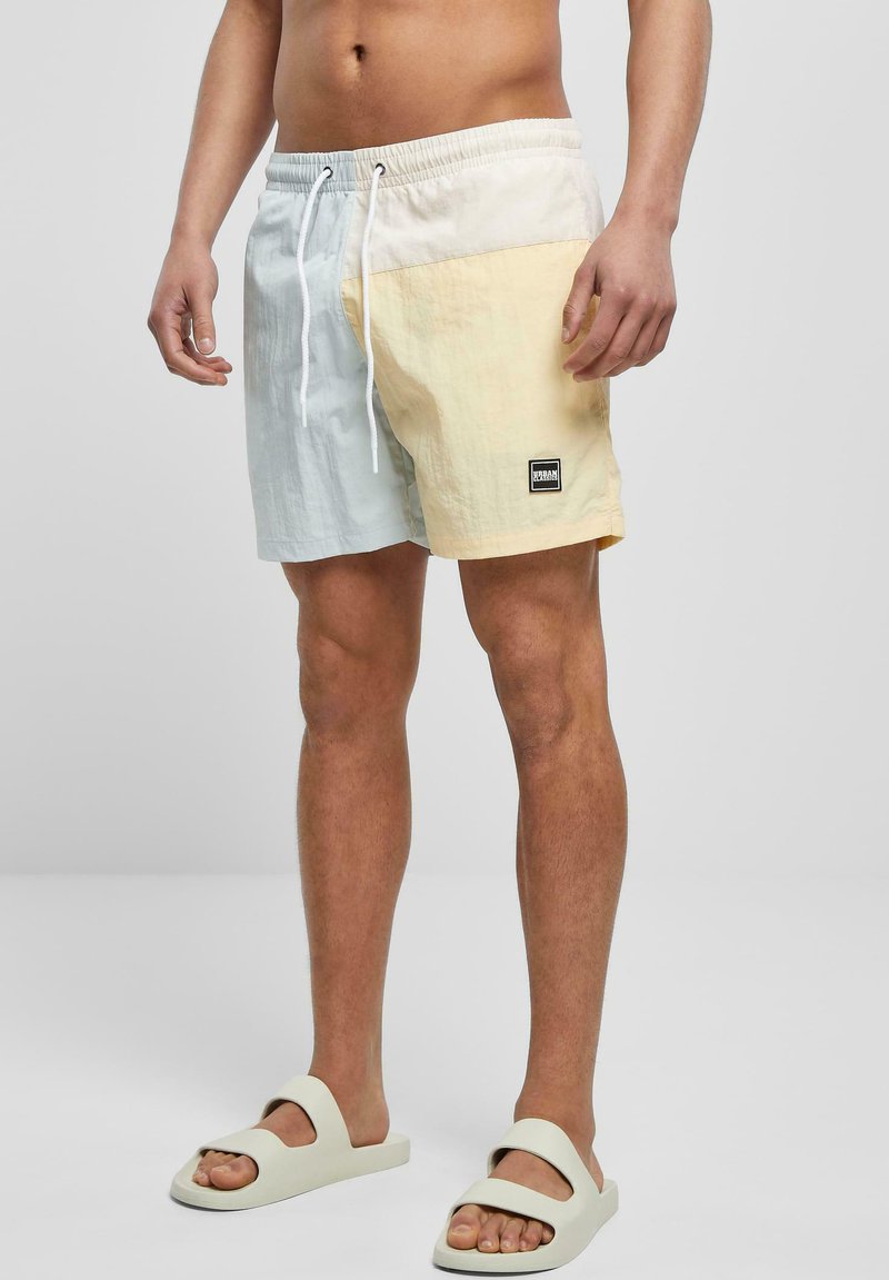 Costume de baie cu un design bicolor în albastru deschis și galben pal, fabricat dintr-un material ușor, cu un brâu elastic și un patch cu logo pe lateral.